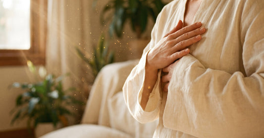 Hands resting at the heart in a soft Qi Gong gesture, golden window light catching dust motes — a moment of nervous system reset